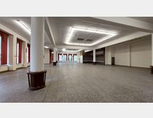 Großer leerer Saal mit gefliesten Böden, weißen Wänden und einer Decke mit flachen Beleuchtungselementen. Die Fensterreihen an der linken Seite sind mit roten Vorhängen versehen, und an der Stirnseite befindet sich eine erhöhte Bühne oder Theke aus dunklem Holz.