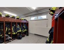 Umkleideraum einer Feuerwehr mit Holzgarderoben, in denen Schutzkleidung und Helme mit reflektierenden Elementen ordentlich verstaut sind. Die Wand hat ein Fenster und darunter befindet sich ein weißer Heizkörper.