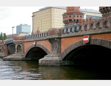 Backsteinbrücke mit zwei steinernen Rundbögen, darüber ein schmiedeeisernes Geländer mit dekorativen Mustern. Im Hintergrund moderne Gebäude und ein großflächiges Gebäude mit gelbem Fassadendesign, während der Fluss im Vordergrund sichtbar ist.