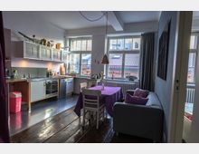 Wohnbereich mit offener Küche und Essbereich in einer Wohnung. Die Küche hat helle Schränke, Edelstahl-Arbeitsflächen und große Fenster, die viel Tageslicht hereinlassen. Im Vordergrund steht ein Esstisch mit lila Tischdecke, daneben ein graues Sofa und Holzboden.