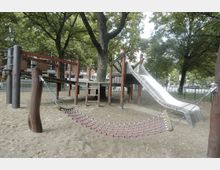 Spielplatz mit Klettergerüst aus Holz, einer angeschlossenen Metallrutsche und einem Hängemattennetz über sandigem Boden. Im Hintergrund sind große Bäume, ein Zaun und parkende Autos zu sehen.