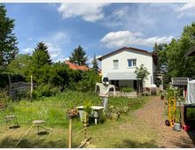 Ein Garten mit einem modernen zweistöckigen weißen Wohnhaus im Hintergrund. Im Vordergrund befinden sich eine Grünfläche, ein selbstgebauter Vogelscheuchenständer, Gartenmöbel und verschiedene Gegenstände wie eine alte Badewanne und ein Schubkarren.