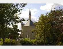 Backstein-Kirche mit spitzem Turm und gotischen Fenstern, umgeben von Bäumen und grünem Laub, bei Tageslicht unter teils bewölktem Himmel. Im Vordergrund ist ein Verkehrszeichen sichtbar.