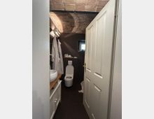 Kleines Badezimmer mit gemauertem Gewölbedecken, dunklen Wand- und Bodenfliesen, einer weißen Tür sowie Waschbecken auf einem Holzunterschrank links und einer Toilette im Hintergrund. Ein kleines Fenster sorgt für natürliches Licht.