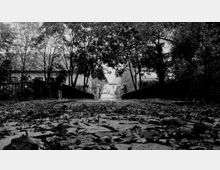 Schwarz-Weiß-Aufnahme eines gepflasterten Weges, der mit Herbstblättern bedeckt ist, flankiert von niedrigen Metallbrücken und umgeben von Bäumen. Im Hintergrund sind Gebäude mit Ziegeldächern und ein schmiedeeisernes Tor zu erkennen.