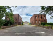 Ziegelgebäude auf beiden Seiten eines asphaltierten Weges, der zu einem größeren historischen Gebäude im Hintergrund führt. Umgeben von Bäumen und grünen Zäunen, unter einem bewölkten Himmel.