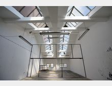 Industriehalle mit weißen Wänden und großen, schrägen Oberlichtern im Dach, die viel Tageslicht hereinlassen. Im Inneren befinden sich schwarze Metallrahmen, hängende Leuchtstofflampen und ein Betonboden.