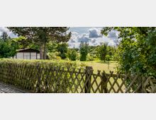 Großer Garten mit Rasenfläche, vereinzelten Bäumen und einer Hecke, im Vordergrund ein Holzzaun. Im Hintergrund ist ein weißes Gartenhaus mit braunen Holzleisten sichtbar, umgeben von dichter Vegetation.