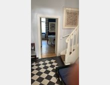 Wohnungsinnenraum mit einem schwarz-weiß karierten Boden im Vordergrund, einer weißen Treppe rechts und einer Türöffnung, die in einen Raum mit Holzboden und einem Tisch führt. An den Wänden hängen gerahmte Bilder, und ein großes Fenster mit gestreiften Vorhängen ist im Hintergrund zu sehen.