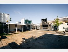 Ein offener Platz mit zusammengesetzten Seecontainern, die als Gebäude dienen, umgeben von Kies- und Pflasterboden. Eine große Lautsprecherinstallation und ein weiß überdachter Bereich sind sichtbar, mit vereinzelt gepflanzten Bäumen im Hintergrund.