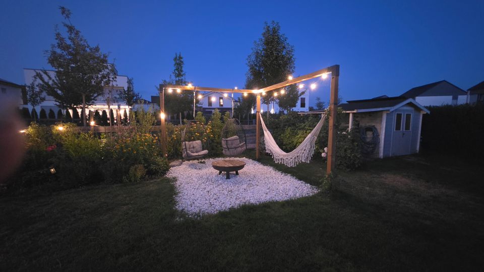 Garten-Lounge im Wohngebiet bei Dämmerung mit Holzpergola und Lichterkette über einer Kiesfläche, darauf eine Hängematte, Sitzmöbel und eine niedrige Feuerstelle. Umgeben von Rasen, Blumenbeeten und Sträuchern, rechts mit kleinem Gartenhaus.