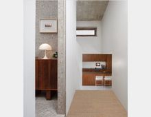 Minimalist interior hallway leading to a compact kitchen with dark wood cabinets, a narrow frosted window, and an exposed concrete ceiling. At left, an alcove with terrazzo walls contains a wooden cabinet topped with a white dome lamp and a framed picture.