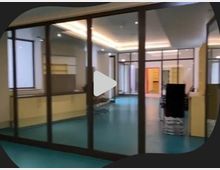 Ein moderner Innenraum mit verglasten Trennwänden, leuchtend türkisfarbenem Boden und einer Decke mit integrierter Beleuchtung. Im Hintergrund sind weitere Räume und offene Durchgänge erkennbar.