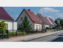 Reihen von Einfamilienhäusern mit Satteldächern und hellem Putz, eingerahmt von Zäunen und begrünter Vorgärten. Eine Bushaltestelle befindet sich am Gehweg entlang der asphaltierten Straße.