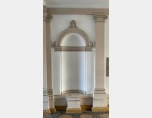 Wandnische in einem Gebäude mit klassizistischen Elementen, umgeben von zwei verzierten Pilastern und einem Bogen mit dekorativem Abschluss. Der Boden ist mit gemusterten Fliesen ausgelegt.