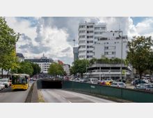Straßenszene in einer Stadt mit einem erhöhten weißen Wohngebäude im Hintergrund, mehreren Autospuren und einem Tunnel im Vordergrund. Auf der linken Seite fährt ein gelber Bus, während Bäume und geparkte Autos die Straße säumen; rechts sind ein Parkhaus und weitere Autos zu sehen.