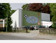 Ansicht eines modernen Gebäudes mit hellgrüner Wand, auf der Weltkarte, Fußball-Symbol und Logos von Fußballverbänden abgebildet sind. Im Vordergrund steht ein offener Parkplatz mit angestellten Autos, umgeben von Bäumen und Holzzäunen.