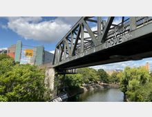 Eine moderne Stahlbrücke mit Graffiti überspannt einen schmalen Kanal, der von grüner Vegetation und Bäumen umgeben ist. Im Hintergrund sind ein Gebäudekomplex mit einem großen gelben BVG-Logo sowie weitere städtische Strukturen zu sehen.