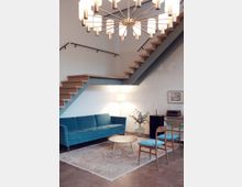 Wohnbereich mit schlichtem, modernem Design: Eine blaue Couch, ein kleiner runder Couchtisch und zwei blaue Stühle stehen auf einem hellen Teppich. Eine Holztreppe mit Metallgeländer führt schräg nach oben, und eine große Deckenleuchte mit mehreren Lichtern dominiert den Raum.