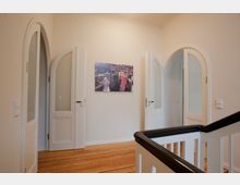 Ein helles Treppenhaus mit weiß gestrichenen Wänden, einem Holzfußboden und einer schwarzen Holztreppengeländer. Drei weiße Türen mit ovalen Glaseinsätzen und Bögen befinden sich an den Wänden, während ein Bild an der zentralen Wand hängt.