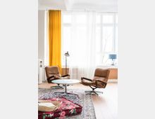 Wohnzimmer mit großen Fenstern, gelben und weißen Vorhängen. Zwei braune Ledersessel stehen um einen runden weißen Tisch, auf einem gemusterten Teppich. Links ist eine Stehlampe, im Vordergrund ein gefaltetes Blumenmusterpolster mit einem Korb darauf.