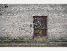 Backsteinmauer mit einer hölzernen Tür, die mit Graffiti bemalt ist. Vor der Mauer befindet sich ein schmaler Gehweg mit Pflastersteinen.