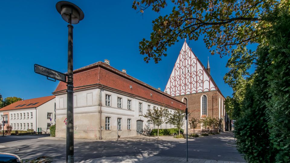 Straßenecke mit einer weiß verputzten, zweigeschossigen Gebäudereihe mit roten Ziegeldächern, daneben eine große Backsteinkirche mit steilem, weiß-rot gemustertem Giebel und hohen Spitzbogenfenstern. Im Vordergrund stehen ein Laternenmast mit Straßenschild sowie Bäume und Grünbewuchs am Straßenrand.