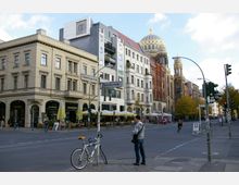 Straßenszene in einer Stadt mit historischen und modernen Gebäuden, darunter die Neue Synagoge mit einer goldenen Kuppel. Im Vordergrund sind Straßenschilder, ein Fahrrad und ein Fußgänger zu sehen, während Cafés mit Außenbereichen am Gehweg liegen.