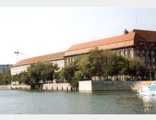 Großes historisches Gebäude mit rotem Satteldach und mehreren Etagen, gelegen an einem Flussufer. Bäume säumen die Uferpromenade, während links im Hintergrund ein Baukran zu sehen ist.
