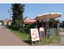 Straßenszene mit einem kleinen Imbiss namens „Feldküche“ neben einem Gehweg; charakteristische Elemente sind ein Sonnenschirm, Werbetafeln mit Tagesangeboten, sowie ein Schild mit der Aufschrift „lecker und reichlich“. Im Hintergrund sind Wohnhäuser mit Gärten zu sehen, und links verläuft eine wenig befahrene Straße.