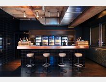 Barbereich mit einer glänzenden, schwarzen Fliesenverkleidung und einer Theke mit Kupferoberfläche. Dahinter befinden sich beleuchtete Kühlschränke mit Getränken, darüber eine Decke mit sichtbaren Lüftungselementen und Spotlichtern.