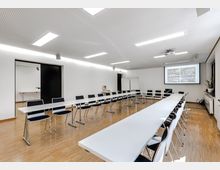 Moderner Seminarraum mit langen, weißen Tischen, schwarzen Stühlen und hellem Holzboden. An der Stirnseite befindet sich eine Leinwand mit Projektor sowie zwei Whiteboards; der Raum hat eine schlichte, weiße Decke mit integrierten Beleuchtungselementen.