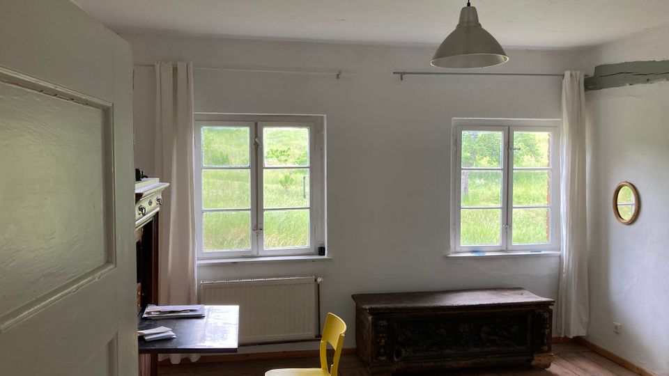 Ein schlicht eingerichteter Raum mit Holzboden und weiß gestrichenen Wänden. Zwei Fenster mit weißen Vorhängen lassen Tageslicht in den Raum, der mit einem schwarzen Tisch, einem gelben Stuhl, einer antiken Holztruhe und einem kleinen runden Spiegel ausgestattet ist.