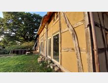 Außenansicht eines Fachwerkhauses mit Lehmwänden, großen Glasfenstern und Holzbalken, umgeben von einer grünen Wiese und Bäumen im Hintergrund. Ein Regenrohr ist am Haus angebracht, und im Vordergrund liegen Backsteine sowie weitere Gegenstände.