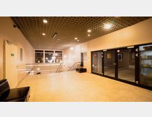 Heller Eingangsbereich mit beigem Fliesenboden, einer schwarzen Ledersitzbank und einer Metallbrüstung. Rechts befinden sich große Glastüren, links eine beige Tür mit Notausgangsschild, und die Decke ist mit einem Gittermuster gestaltet.