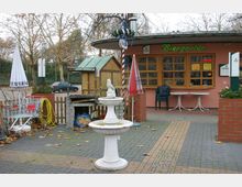 Außenbereich eines Biergartens mit rosa Fassade, Holztüren und -fenstern. Vor dem Gebäude stehen ein weißer Zierbrunnen, Tische mit Stühlen sowie Sonnenschirme, umgeben von gepflastertem Boden und kahlen Bäumen.