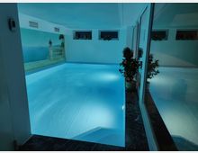 Innenansicht eines beleuchteten, rechteckigen Indoor-Schwimmbeckens mit hellblauem Wasser. An den Wänden befinden sich ein Wandgemälde mit Meeresmotiv, kleine Fenster und dekorative Pflanzen in Blumentöpfen, die den Raum ergänzen.