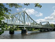Grüne Stahlbrücke mit bogenförmigen Verstrebungen, die über einen Fluss führt, umgeben von Bäumen und klarem Himmel mit weißen Wolken. Die Brücke steht auf massiven Steinpfeilern im Wasser.