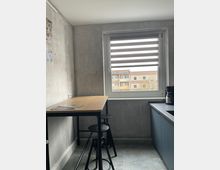 Kleine Küche mit Betonoptik-Wänden, einem hohen Esstisch aus Holz mit zwei schwarzen Barhockern, einem Fenster mit gestreifter Jalousie und blauen Küchenschränken. Im Hintergrund sind Wohnhäuser durch das Fenster sichtbar.