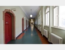Ein heller, langer Flur mit cremefarbenen Wänden und einem dunkelgrünen Boden, gesäumt von roten Holztüren mit Steinrahmen auf der linken Seite und großen, bogenförmigen Fenstern sowie Heizkörpern auf der rechten Seite. Die Decke ist mit hängenden Kugelleuchten beleuchtet.