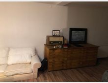 Wohnzimmer mit einer cremefarbenen Couch links und einer großen, hölzernen Kommode rechts. Auf der Kommode stehen ein Vintage-Radio, ein gerahmtes Bild sowie Bücher und andere kleine Gegenstände. Der Boden besteht aus Holzlaminat, und die Wände sind schlicht weiß.