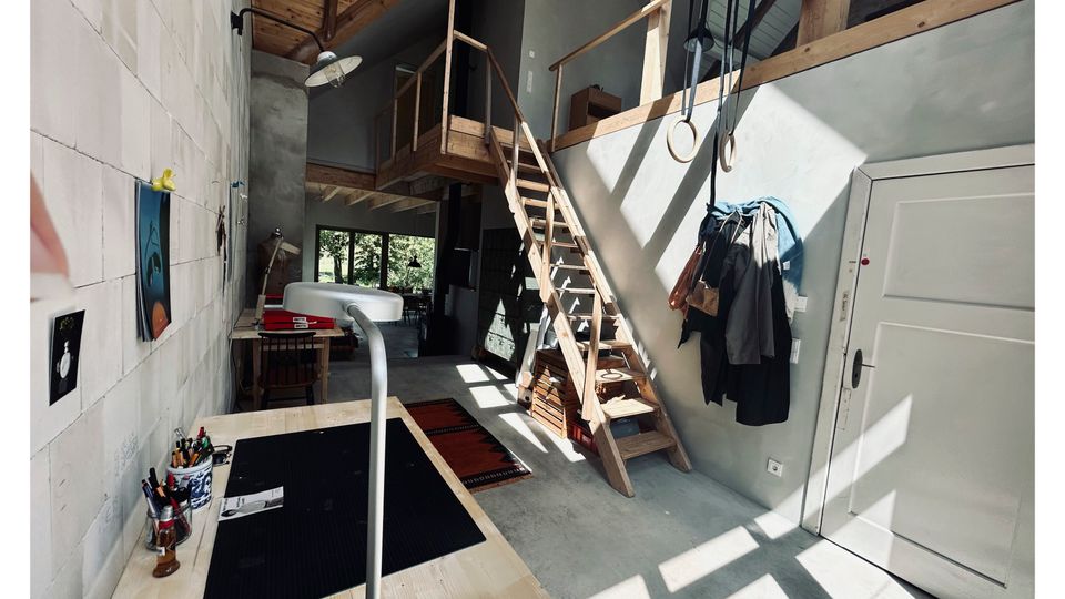 Blick von oben in einen hohen, hellen Innenraum mit Sichtbetonwänden, großen Fenstern und eingezogenem Holz-Zwischengeschoss mit Treppe und Geländer. Unten sind ein Fliesenboden sowie eine Werkbank mit Werkzeugen und Materialien zu sehen, durch die Fenster fällt starkes Sonnenlicht mit Schattenmustern.