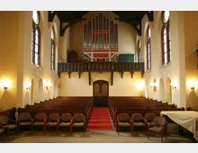 Innenraum einer Kirche mit Holzbänken und einer zentralen Sitzreihe, die auf einen roten Teppich ausgerichtet ist. Über der Eingangstür befindet sich eine Balustrade mit einer Orgel, umgeben von hell gestrichenen Wänden und hohen bunten Fenstern.