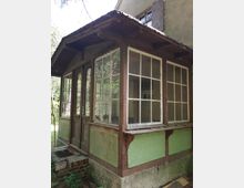 Ein verlassener Eingangsanbau mit großflächigen, verglasten Fenstern, braunen Holzrahmen und grüner Putzfassade. Das Gebäude zeigt erkennbare Alters- und Verwitterungsspuren, umgeben von Waldvegetation.