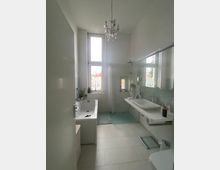 Geräumiges Badezimmer mit einer Badewanne links, einer großen Fensterfront im Hintergrund, einer Dusche mit Duschvorhang daneben und einem langen Waschbecken mit Spiegel rechts. Der Raum ist in hellen Farben gehalten und wird von einem Kronleuchter beleuchtet.