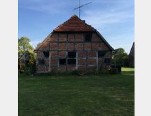Fachwerkhaus mit rotem Ziegeldach in ländlicher Umgebung, umgeben von einer Rasenfläche und Blumenbeeten. Im Hintergrund sind weitere Gebäude und Bäume zu erkennen.