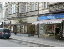 Straßenszene mit einer Reihe von Geschäften in einem Altbau. Zu sehen ist ein Uhrengeschäft mit gewölbten Fenstern, daneben ein Lederwarengeschäft und ein Schuhgeschäft mit einer Markise und ausgestellten Schuhen auf einem Ständer vor dem Laden.