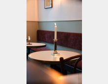 Kleiner Tisch in einem Café mit einer brennenden Kerze in einem silbernen Kerzenhalter. Im Hintergrund sind Sitzbänke mit rotem Polster und eine pastellfarbene Wand mit einem gerahmten Bild sichtbar.