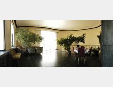 Wohnzimmer mit cremefarbenen Wänden und dunklem Fliesenboden, ausgestattet mit mehreren großen Zimmerpflanzen, einer Sitzecke mit Sofas und Sesseln sowie einem Esstisch mit roten Stühlen. Eine große Glasfront mit weißen Vorhängen lässt helles Tageslicht in den Raum.