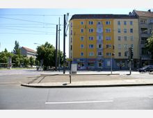 Straßenszene mit einer gelben Wohn- und Geschäftsgebäudefassade, im Erdgeschoss befinden sich mehrere kleine Geschäfte. Im Vordergrund sind Straßenbahngleise, eine Straßenkreuzung und Verkehrsschilder zu sehen, umgeben von Bäumen und einer weiteren Gebäudezeile im Hintergrund.